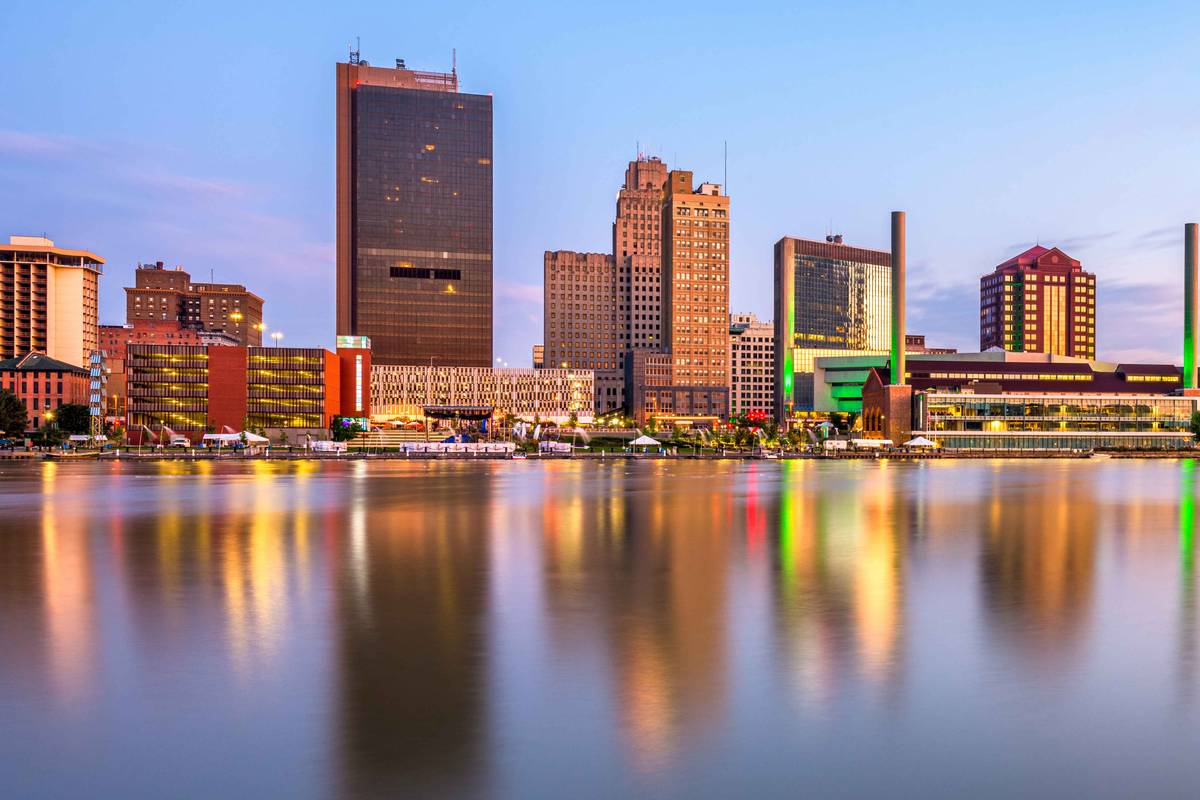 The Toledo skyline at dusk mirrored in the Maumee River, with a dark glass tower at the center and waterfront lights reflecting across the water.